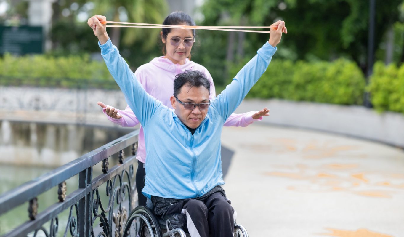 A man in a wheelchair exercising, supported by his partner during a resistance, strengthening and cardio workout routine. This image highlights the themes of fitness, motivation, and perseverance.